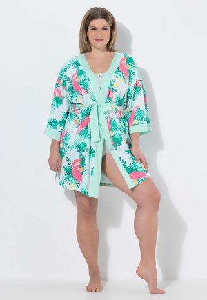 Femme portant un court peignoir vert menthe avec des motifs d'oiseaux roses et de feuilles vertes, noué à la taille, debout pieds nus sur un pied.