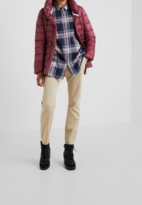 Bordeaux-farbene gepolsterte Jacke, karierte Hemd in Marineblau und Weiß, beige Chinos und schwarze Schnürstiefel. Glatte Textur und lässiges Design.
