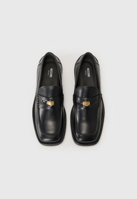 Schwarze Lederloafers mit quadratischer Schuhspitze, gestickten Details und einem goldenen Emblem auf dem Riemen. Glatte Textur und elegante Silhouette.