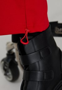 Black leather boot with two adjustable straps, paired with red adjustable pant cuffs featuring red drawstring accents.
