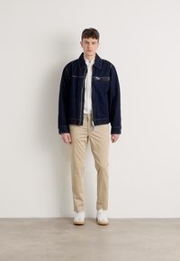 Tumši džinsi jaka ar priekšējo rāvējslēdzēju, zem tās baltā krekla ar pogām, gaiši brūni chino auduma bikses un baltas sporta apavas uz neitrāla fona.