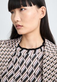 Strickoberteil mit einem schwarzen, rosa und weißen Rautenmuster, das einen gewellten Halsauschnitt hat. Begleitet von einem gemusterten Blazer in passenden Farben.