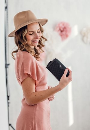 Black zippered wallet with patterned texture held by a person in a light pink dress. The wallet has a distinctive logo on one side.