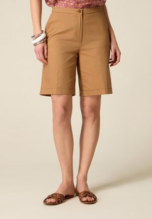 Vrouw draagt beige, knielange, op maat gemaakte shorts, bruine gevlochten sandalen, zilveren en witte armbanden en een roze top met patroon, staand tegen een beige achtergrond.