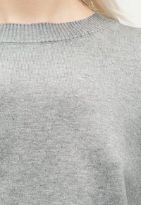 Κοντινό πλάνο ενός ατόμου που φοράει ανοιχτό γκρι ριμπ πουλόβερ με στρογγυλή λαιμόκοψη, εμφανίζοντας την υφή της πλέξης και την περιοχή του άνω στήθους.