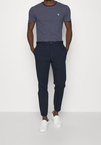 Pruhované námořnické tričko s kulatým výstřihem, tmavé kalhoty a bílé klouzací boty. Kalhoty mají přiléhavý střih a boční kapsy.