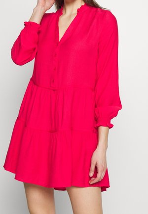 Kobieta w jasnoróżowej, długorękawowej sukience z falbankami i zapinanej na guziki z przodu, stojąca na jednolitym jasnym tle.