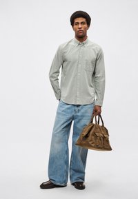 Svetlo zelena srajca z gumbi in žepom na prsih, kombinirana z širokimi modrimi jeans hlačami in rjavo usnjeno torbo, ki ima stranske žepe in ročaje.