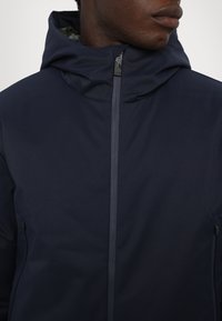 Giacca con cappuccio di colore blu navy con una finitura liscia e opaca. Presenta una cerniera frontale completa, due tasche laterali con zip e un colletto alto.