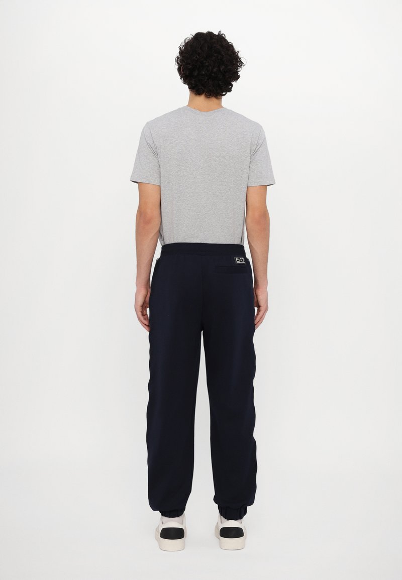 T-shirt grigio a maniche corte abbinato a pantaloni della tuta blu navy con vita elasticizzata e caviglie elastiche. Sneakers bianche con accenti neri.