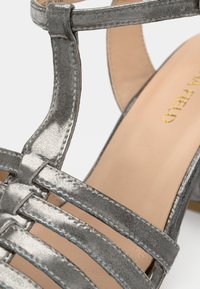 Metāliskas sudraba sieviešu sandales ar siksniņām, gludu tekstūru, atvērtu papēdi un zemu kvadrātveida papēdi. Redzama bēša zolīte.