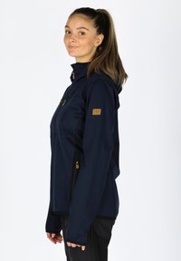 Marineblaue Softshelljacke mit tailliertem Design, Reißverschlusstaschen und Kapuze. Verfügt über braune Logoakzente auf Ärmel und Brust. Glatte Textur.