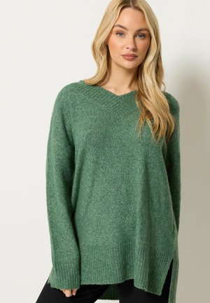 Blonde vrouw draagt een losse, groene V-hals gebreide trui met lange mouwen en zijsplitsen, staand tegen een effen lichte achtergrond.