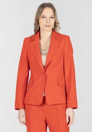 Kobieta o jasnobrązowych włosach ubrana w jasnopomarańczową dopasowaną marynarkę i spodnie w tym samym kolorze, na beżowej bluzce, stoi na jednolitym tle.