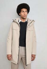 Parka beige claro con capucha, cierre frontal con cremallera y botones, dos bolsillos en el pecho y detalles en negro. Llevado sobre un suéter oscuro y una camisa.