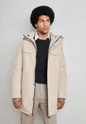 Parka beige claro con capucha, cierre frontal con cremallera y botones, dos bolsillos en el pecho y detalles en negro. Llevado sobre un suéter oscuro y una camisa.
