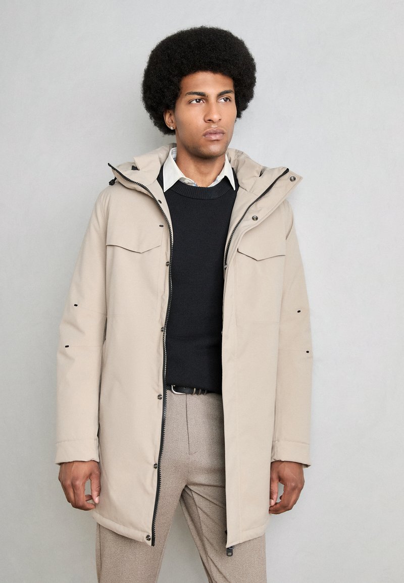 Parka beige claro con capucha, cierre frontal con cremallera y botones, dos bolsillos en el pecho y detalles en negro. Llevado sobre un suéter oscuro y una camisa.