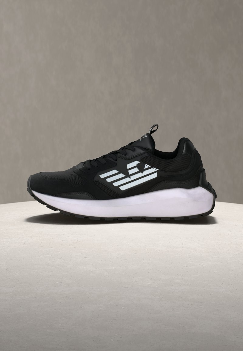 Μαύρο αθλητικό παπούτσι με λευκό στυλιζαρισμένο λογότυπο φτερού στο πλάι και χοντρή λευκή σόλα, τοποθετημένο σε ανοιχτή επιφάνεια με γκρι φόντο.