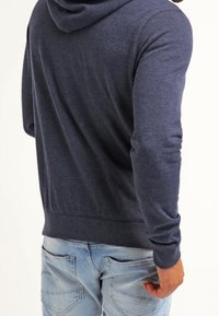 Man i en marinblå huvtröja och ljusblå jeans, sedd bakifrån med en hand synlig.