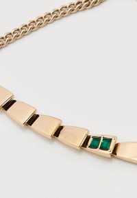 Χρυσό βραχιόλι με ορθογώνιους κρίκους, δύο πράσινες ορθογώνιες πολύτιμες πέτρες και ένα διακριτικό ανάγλυφο λογότυπο κροκοδείλου σε έναν κρίκο.