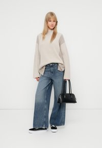 Pulover bej cu riduri, cu mâneci lungi, asortat cu blugi largi de culoare albastră. Geantă neagră și adidași alb-negru ca accesorii.