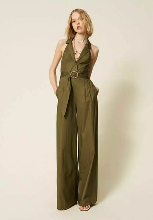 Donna che indossa una tuta senza maniche color oliva con cintura, gambe larghe e scollatura profonda a V, in piedi con le mani nelle tasche davanti a uno sfondo semplice.