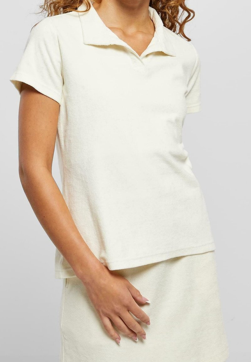 Femme portant un polo crème à manches courtes et une jupe assortie, debout, main posée sur la jupe, devant un fond uni.