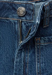 Denim hlače imajo temno modro barvo z vidnim zadrgo in gumbom, svetlo rjavimi šivi in teksturirano tkanino s rahlim bledenjem.