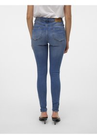 Femme portant un jean skinny bleu taille haute et des mules noires à talons, debout de dos sur un fond uni.