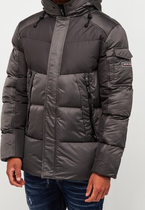 Homme portant une veste d'hiver matelassée grise avec capuche, fermeture éclair devant et poches, et une petite écusson sur la manche supérieure.