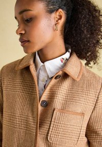 Tan checkered jacket with a pronounced collar, featuring brown trim and a button, paired with a white shirt showcasing a colorful pattern.