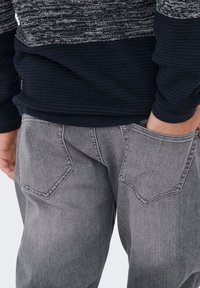 Vaqueros de mezclilla gris con una textura ligeramente descolorida, que cuentan con dos grandes bolsillos traseros angulares y un ajuste casual. Suéter texturizado azul marino oscuro encima.