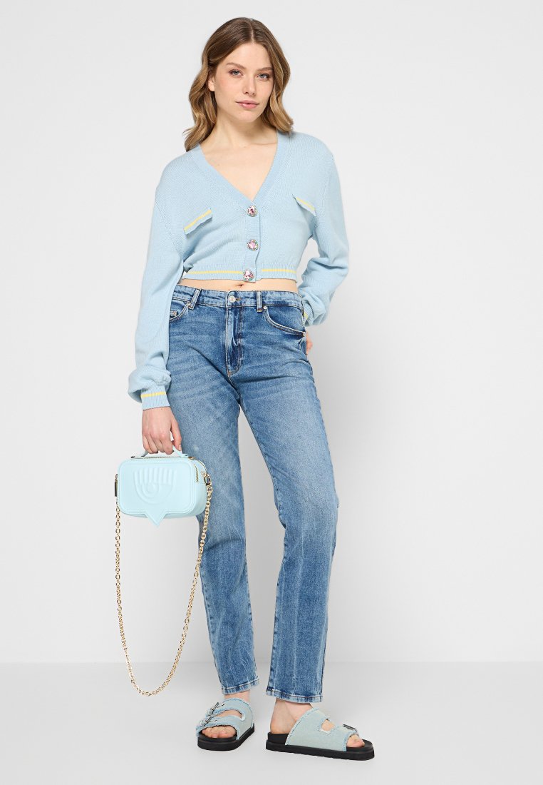 Vrouw in een lichtblauwe cropped cardigan, hoge blauwe jeans, sandalen, met een bijpassende lichtblauwe handtas met een gouden schouderband.