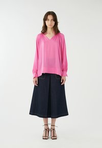 Blusa rosa a maniche lunghe con scollo a V e spalle arricciate, abbinata a una gonna in denim scuro lunga fino alla caviglia con cuciture gialle. Sandali neri con strappi.
