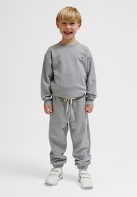 Grauer Pullover mit Rippbündchen, passende graue Jogginghose mit elastischem Bund und Kordelzug. Schwarzes Herz-Logo auf der linken Brust. Weiße Turnschuhe.