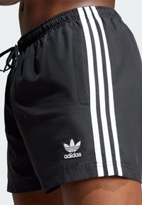 Sorte svømmebukser laget av glatt stoff, med hvite sidestriper og en liten hvit logoaksent. Elastisk midjebånd med snor.