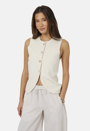 Donna che indossa una maglia crema senza maniche con bottoni e pantaloni chiari a righe con coulisse, che posa con una mano in tasca su sfondo bianco.