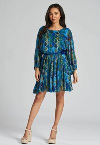 Vestido de manga corta con un patrón geométrico en azul, verde y dorado, falda plisada y un cinturón a juego. La modelo lleva sandalias de plataforma en color beige.