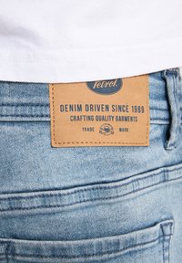 Jeans in denim con etichetta in pelle marrone chiaro con testo blu embossato che recita "Denim Driven Since 1989" e "Crafting Quality Garments."