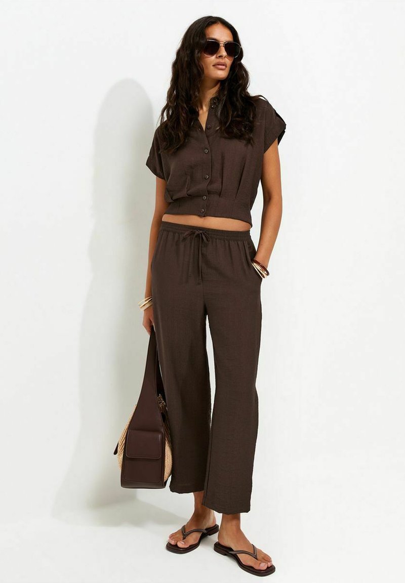 Woman wearing dark brown buttoned short-sleeve top and matching pants, carrying a bag, wearing sunglasses, bracelets, and flip-flop sandals.