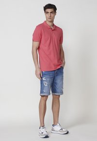 Rdeča polo majica s kragnico, ujemajoča se z raztrganimi modrimi denim kratkimi hlačami. Bele športne superge z sivo in mornarsko modro akcentacijo. Preprost, sproščen videz.