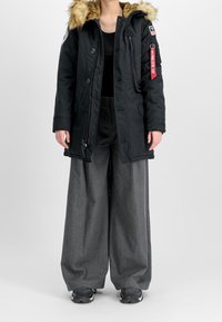 Parka noire avec capuche en fausse fourrure, fermeture zippée et à boutons, poches latérales, et un écusson sur la manche. Associée à un pantalon gris à jambes larges.