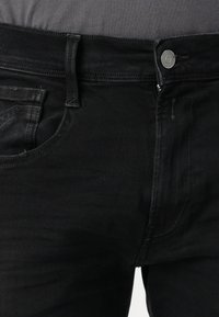 Section avant d'un jean en denim noir avec fermeture par bouton, passants de ceinture, et une chemise grise partiellement visible rentrée à l'intérieur.
