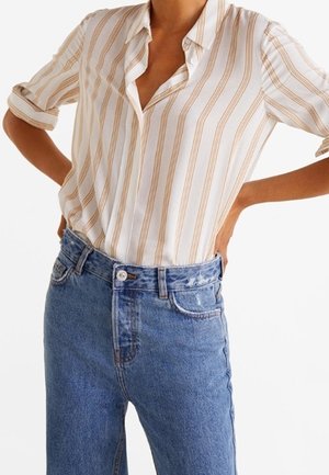 Kobieta w białej koszuli zapinanej na guziki z beżowymi pionowymi paskami i niebieskich dżinsach z wysokim stanem, trzymająca ręce na biodrach.