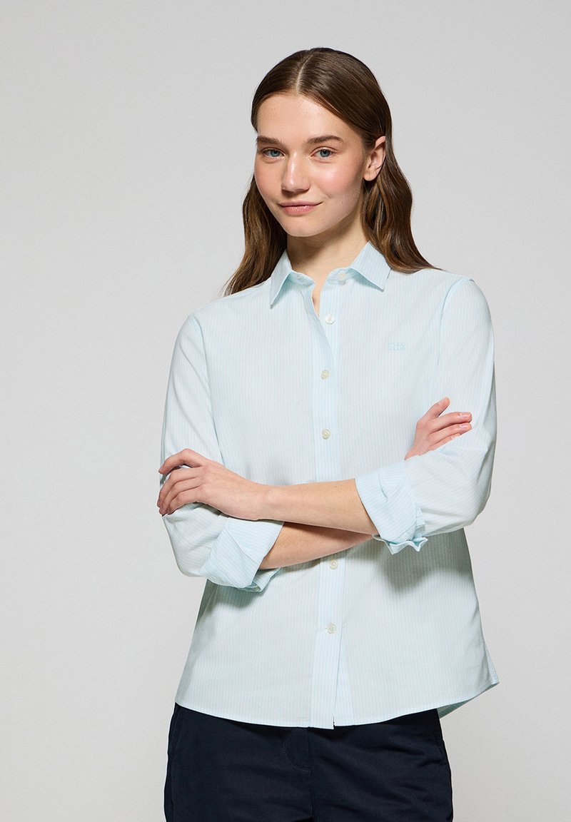 Camicia a righe di colore azzurro chiaro con colletto, maniche lunghe arrotolate e chiusura con bottoni. Realizzata in materiale morbido e dal design a vestibilità regolare.