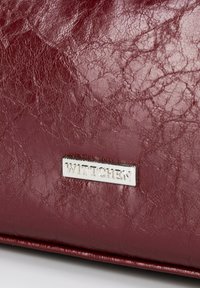 Bordowa skórzana torebka o teksturowanym wykończeniu, z metalową płytką logo "Wittchen" na przodzie, służącą do identyfikacji marki.