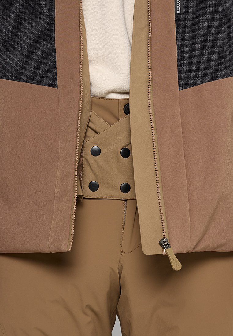 Gros plan sur une veste beige avec une fermeture éclair ouverte, montrant un pantalon marron avec un panneau de taille ajustable à boutons et une chemise blanche en dessous.