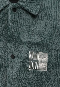 Πράσινο φλις σακάκι με κουμπιά και υφή στην επιφάνεια, με μια μαύρη και λευκή ετικέτα που γράφει "SALINITY NOW BILLABONG".