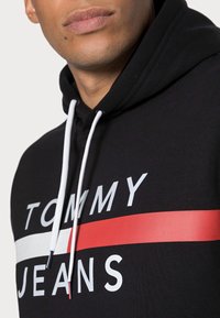 Černá mikina s bílými šňůrkami, která má na hrudi bílý nápis "TOMMY" a "JEANS" a červený pruh. Hladká struktura látky.