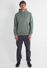 Sweatshirt à capuche verte en tissu doux, avec une poche kangourou à l'avant et un design de logo. Associé à un pantalon bleu foncé et des chaussures noires.